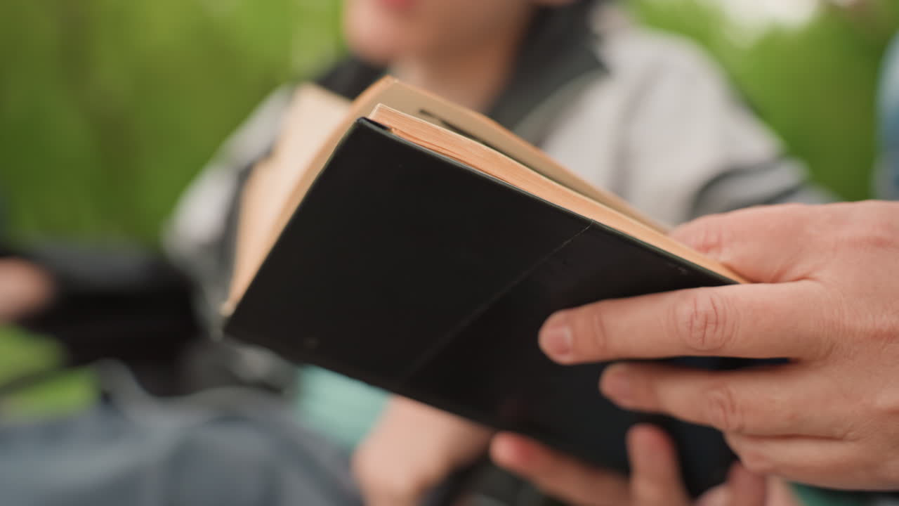Hands Holding Small Black Book In Park Setting With Shallow Depth Of Field Reader Flips Pages Amid Green Blurred Background, Relaxed Posture And Calm Atmosphere, Vintage Paper Texture And Quiet
