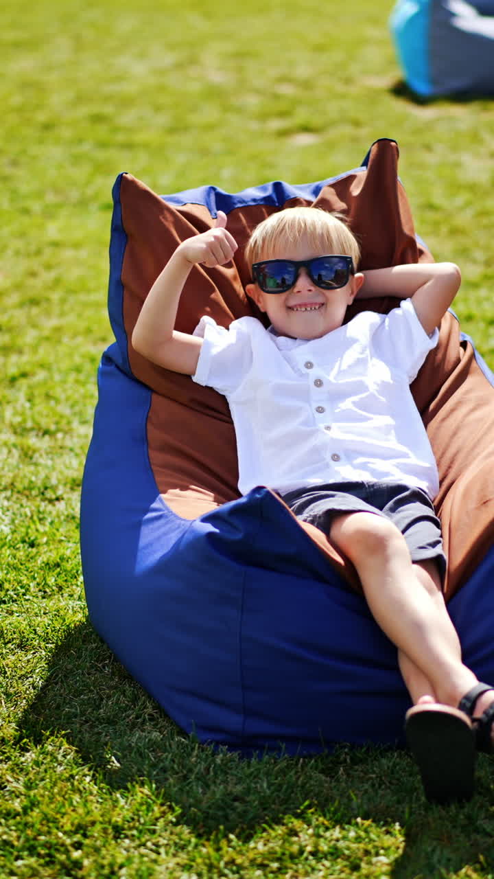 Smiling blond little boy enjoying his time in bean bag chair outdoors. Child in summer clothes and sunglasses relaxing and sunbathing. Vertical video