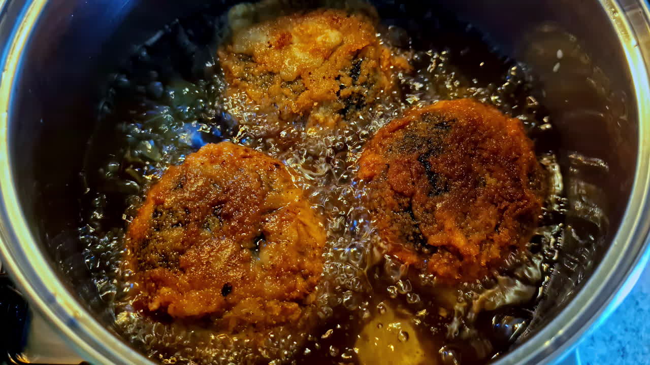 An extreme close-up shot shows three homemade meat patties, similar to Latvian kotletes, deep-frying to a golden-brown crisp in a pot of hot, bubbling, and sizzling cooking oil