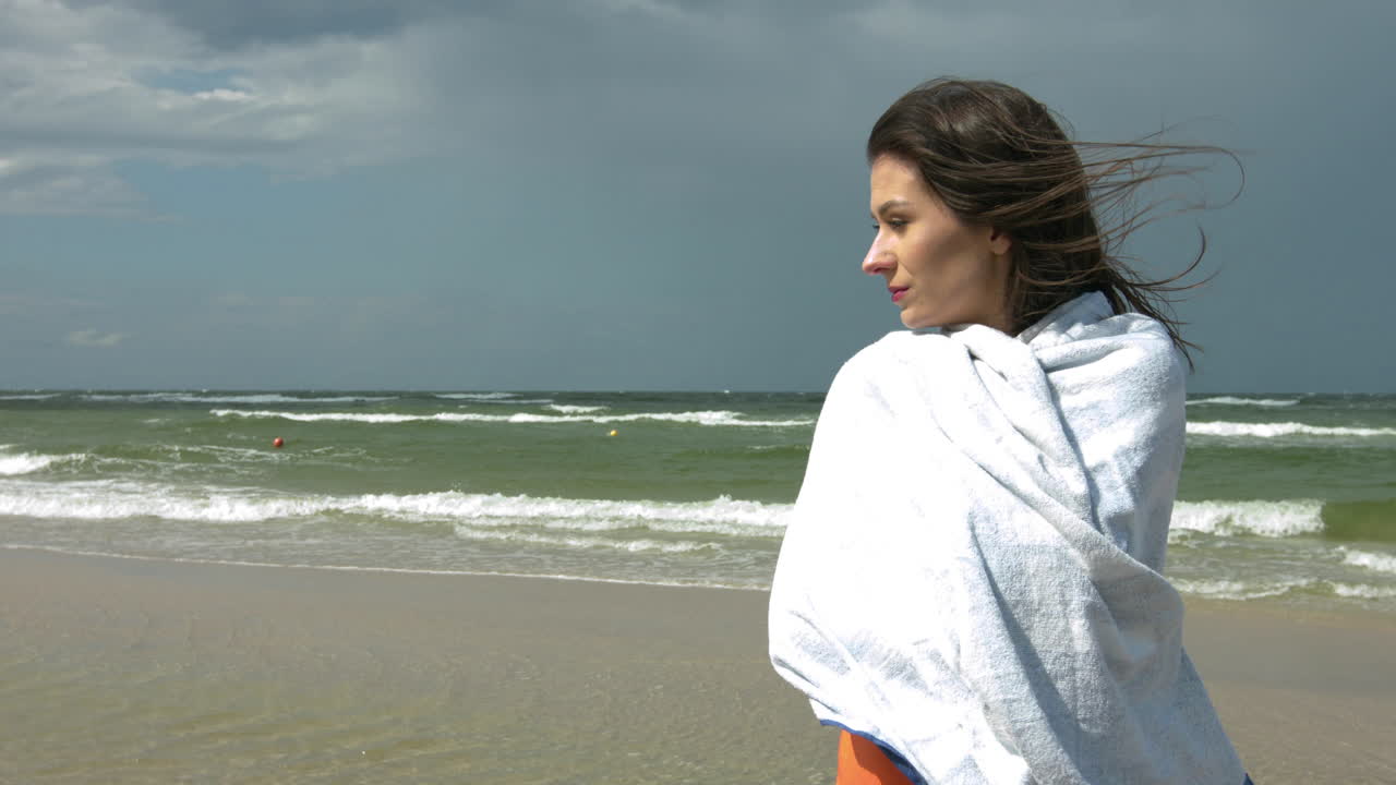 Young woman standing on the seashore and swimsuit covered with a towel on a windy summer day,