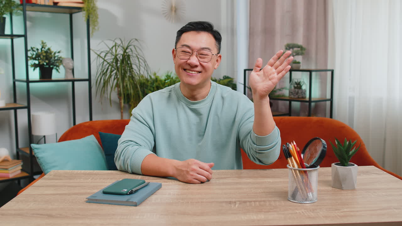 Asian man smiling friendly at camera waving hands gesturing hello hi greeting at home on sofa