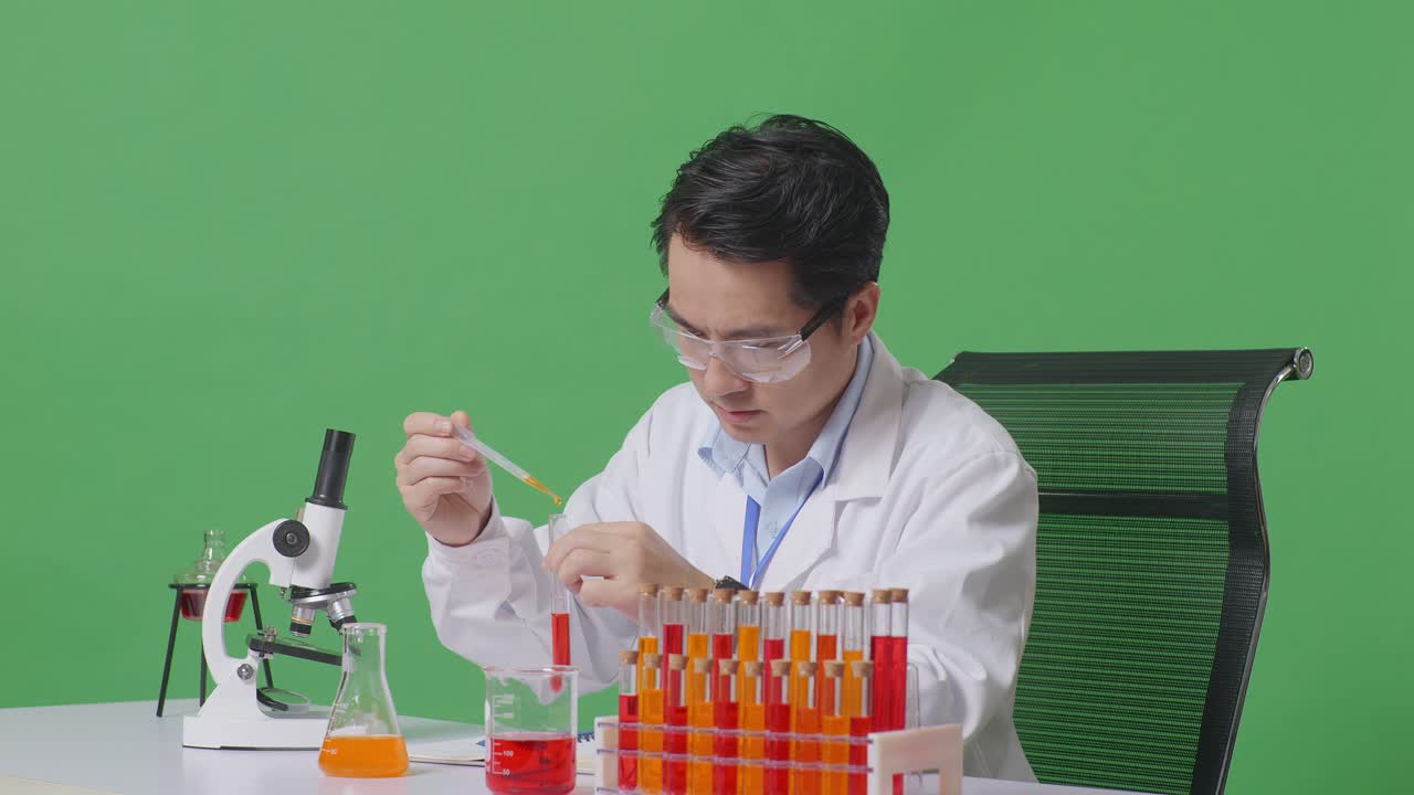 Side View Of Asian Man Scientist Making Experiment With Test Tube And Writing Into Notebook While Working On The Table With Microscope In The Green Screen Background Laboratory