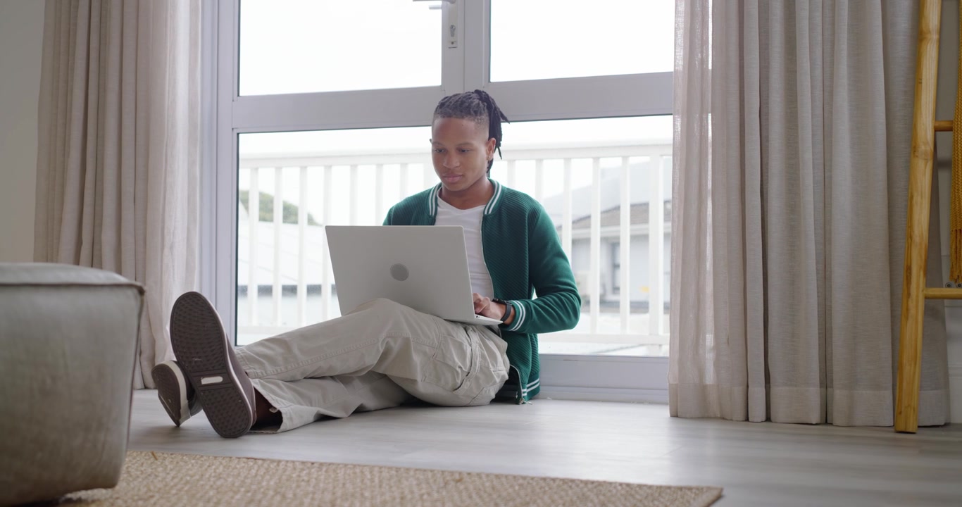 African American man sitting on floor by glass door after settling, typing on laptop and studying
