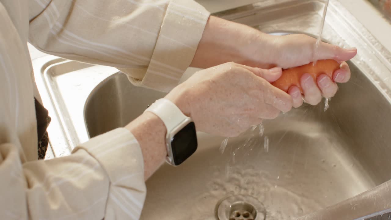 Voksen kvinde ved vasken, vandhanen kører, skyller og skrubber en gulerod, fjerner snavs med et smartwatch.