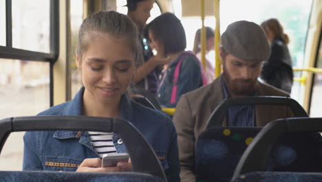 Junge-Frau-Sitzt-In-Einem-Bus,-Während-Sie-Ein-Smartphone-Benutzt,-Dahinter-Ist-Ein-Mann-In-Einer-Baskenmütze,-Der-Auf-Das-Smartphone-Und-Die-Stehenden-Personen-Blickt