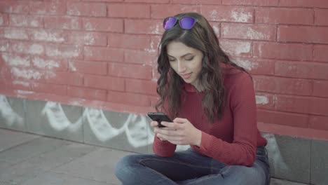 niña sonriente enviando mensajes de texto en un teléfono inteligente con una pared de ladrillos en el fondo