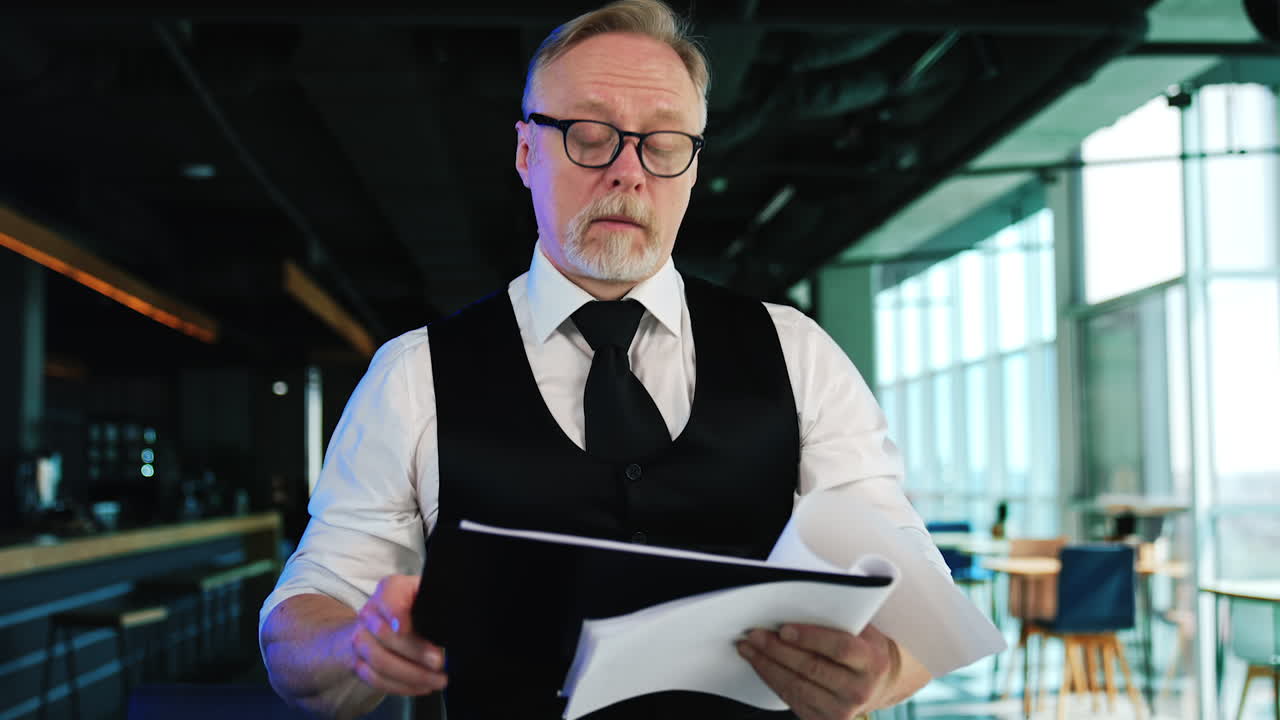 Focused male businessman wearing white shirt and black vest holding papers. Man checks the papers and smiles.