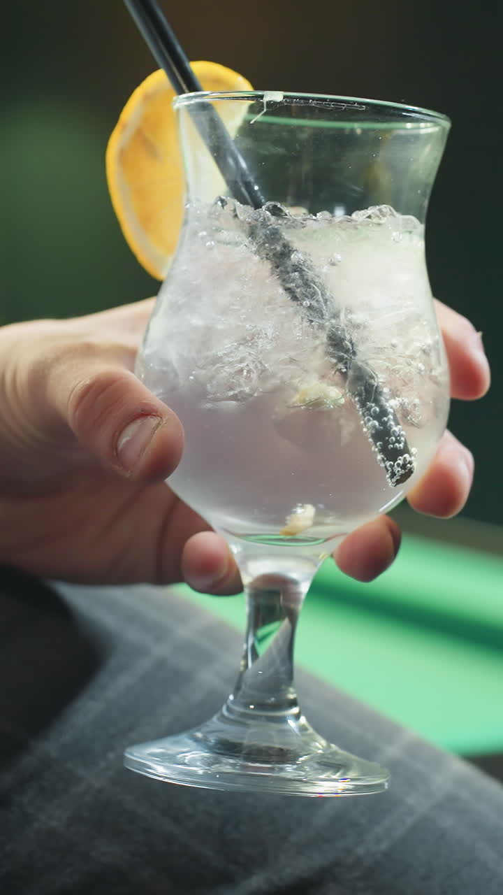 primer plano de una persona, cara no visible, con camisa blanca y pantalones grises, sosteniendo un vaso de bebida de limón con paja negra. cubos de hielo y rebanada de limón agregan detalles refrescantes en un entorno elegante