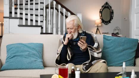 sick senior man sitting on sofa and coughing while typing on smartphone