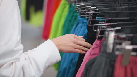 mujer joven navegando por un estante de ropa de colores en una tienda minorista, inspeccionando suavemente prendas de vestir vibrantes que incluyen verde, azul, rosa y negro, entorno brillantemente iluminado