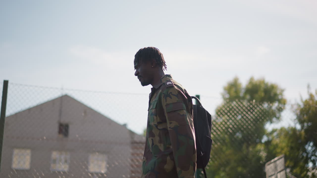 Black Cadet Walking Across Fenced Court With Backpack And Camouflage Jacket Under Clear Sun. Determined Silhouette And Steady Stride Show Training Focus In Suburban Environment. LowRise Buildings