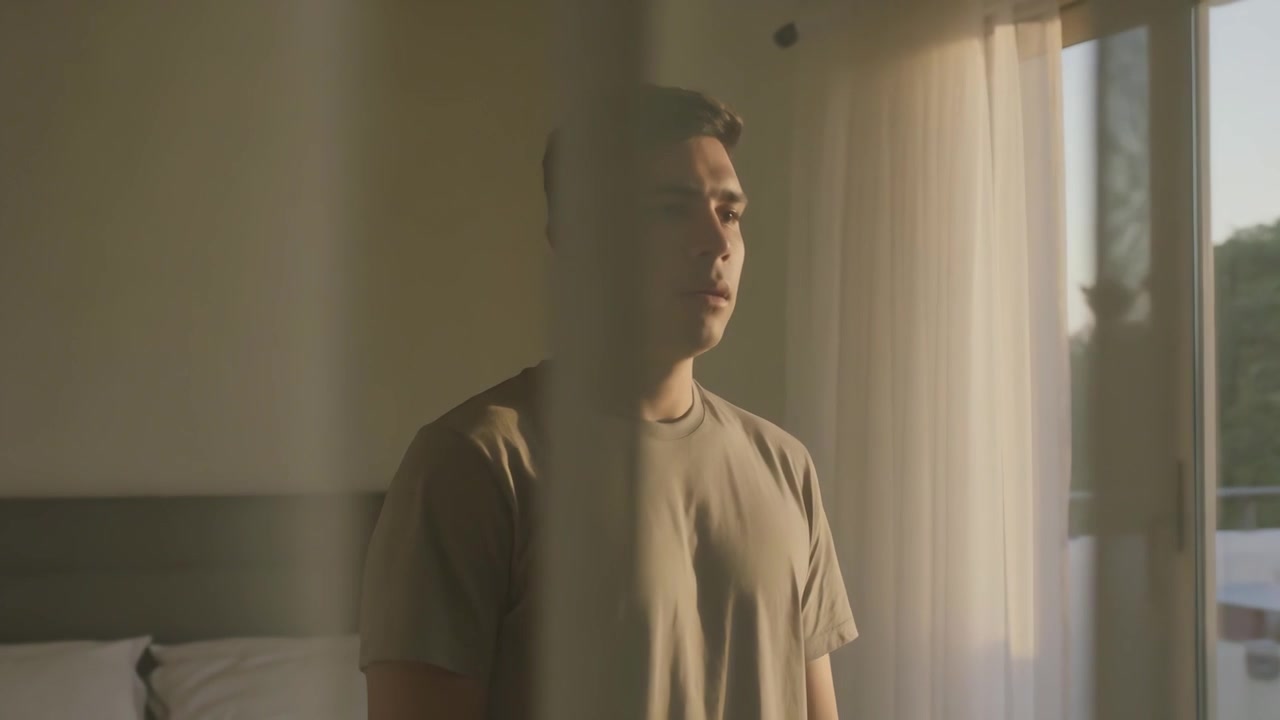 Warm sunlight drawing man in tee turning toward sliding glass door in bedroom, with bed, curtains