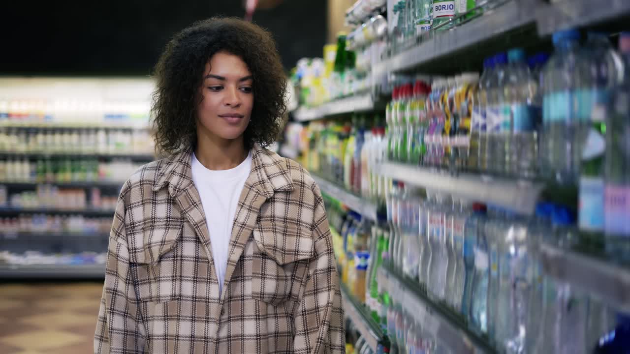mujer negra haciendo compras de comestibles en el supermercado, tomando una botella de agua del estante