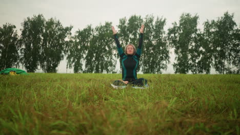 mujer de traje verde y negro sentada en una alfombra de yoga con las piernas cruzadas, las manos levantadas por encima de la cabeza, practicando yoga al aire libre en un campo cubierto de hierba con botas amarillas y tela verde visible en el fondo