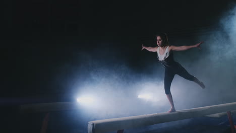 the girl performs a trick on a log in backlight and slow motion in sports gymnastic clothing. smoke and blue. jump and spin on the balance beam