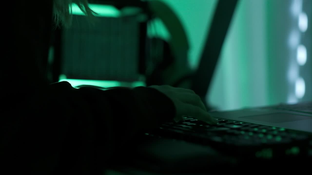 una hackergirl criminal con capucha negra que usa una computadora portátil y computadoras grandes para hackear el sistema de red desde su oscuro cuarto de neón hacker