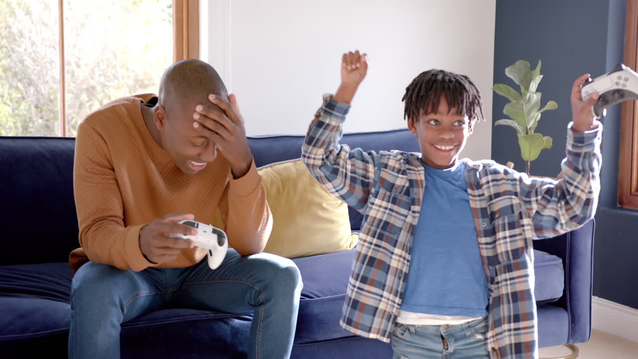 Happy african american father and son playing video games at home, slow motion