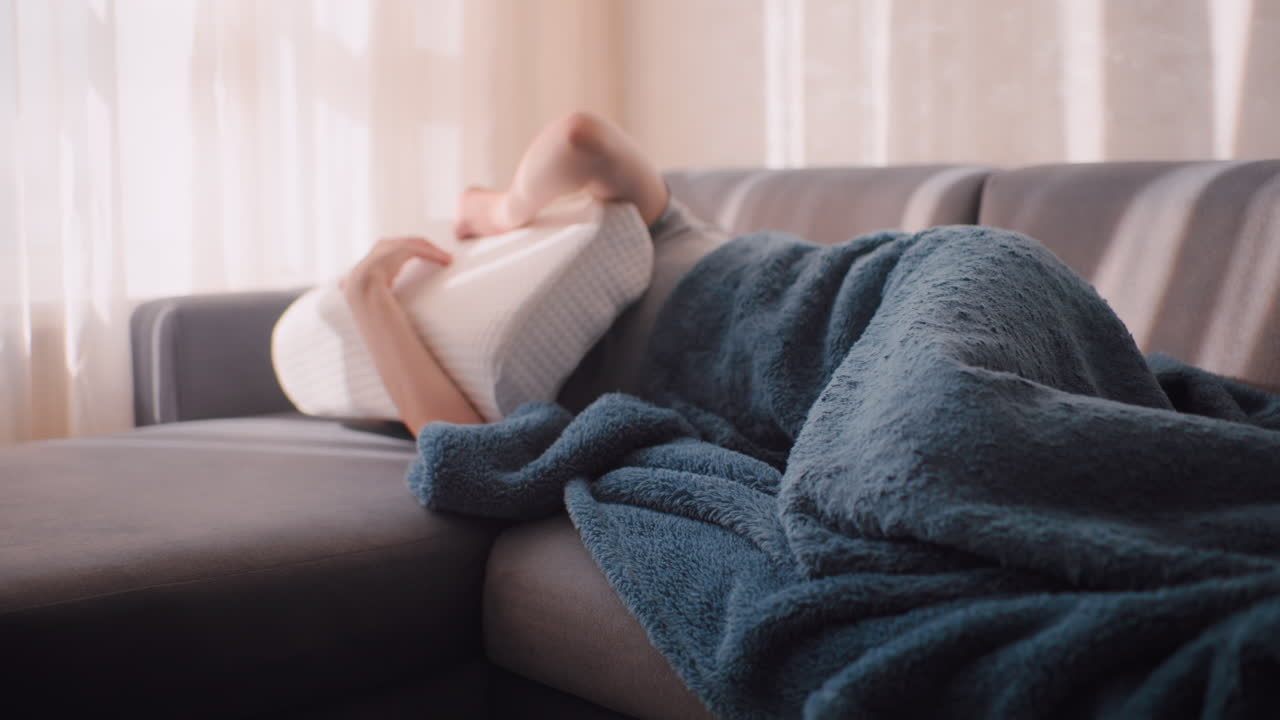 House resident lying on soft couch covers face with fluffy pillow to block bright daylight entering through curtain, resting under blue blanket in quiet room as warm sunbeam fills peaceful interior