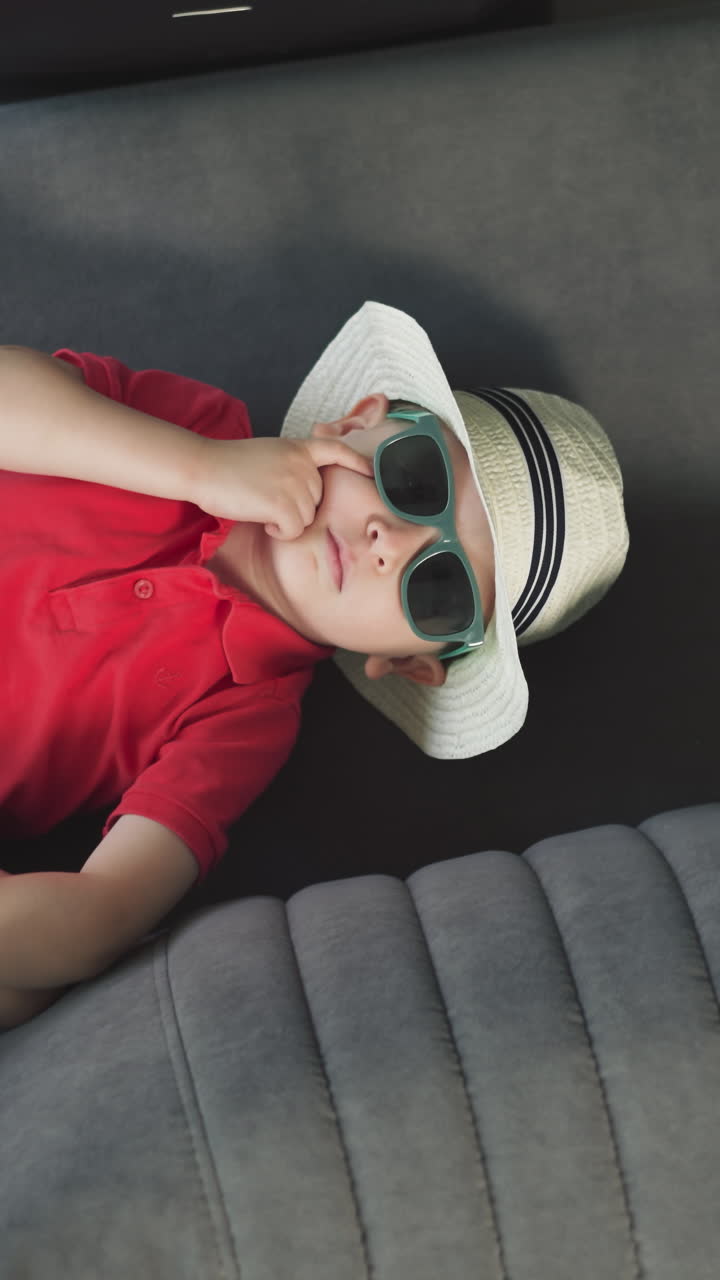 elegante niño pequeño yace en el cómodo sofá en la habitación de la suite del hotel. lindo niño en gafas de sol y sombrero de paja disfruta de las vacaciones vista superior de cerca