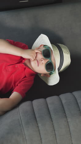 stylish little boy lies on comfortable couch in hotel suite room. cute toddler in sunglasses and straw hat enjoys vacation upper close view