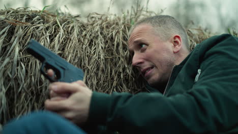 a close view of a man in a green jacket lying in a ditch against dry grass, holding a pistol with a tense expression, and looking around