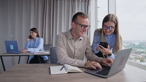 colleagues wearing glasses laughing looking at laptop and phone. female coworker sitting at table behind and talking on the phone.