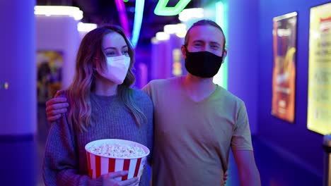 una joven pareja caucásica feliz con máscara va al cine, caminando por el pasillo con un cubo de palomitas de maíz