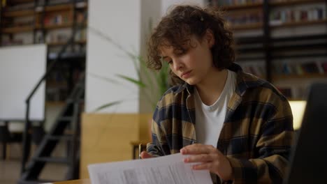 A-girl-student-with-piercings-with-curly-hair-in-a-checkered-shirt-sits-at-a-table-and-reads-protocols-for-work-in-the-university-library