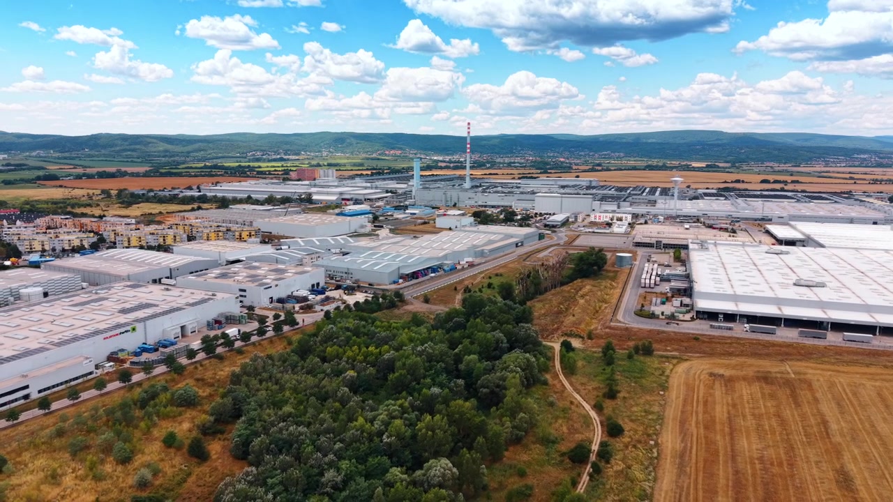 Industridistrikt med varehus og skogkledd forgrunn. Hvite fabrikkhaller, veier, lasteområder, skorsteiner, boligblokker og jordbruksland fyller den brede flyfotoen