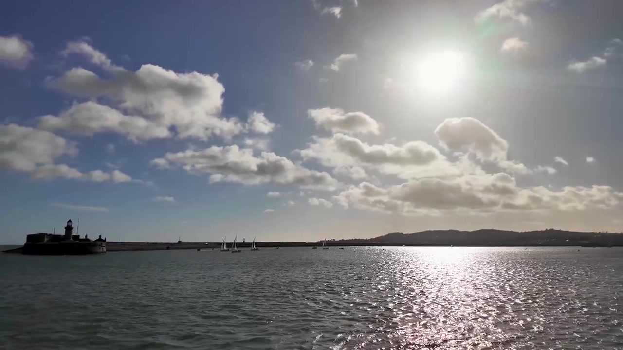 Camminando verso il lungomare a Dún Laoghaire, Irlanda, con vista su barche ormeggiate, la Baia di Dublino e l'iconico Faro di East Pier sotto cieli costieri luminosi.