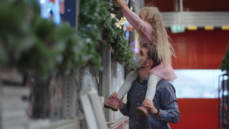 father and daughter sitting on their shoulders in a shopping mall choose a garland for the house and christmas tree for christmas during the pandemic in slow motion.