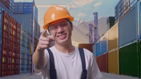 close up de hombre asiático trabajador con gafas de protección y casco de seguridad sonriendo y tocando su pecho luego apuntando a usted mientras está de pie en el depósito de contenedores
