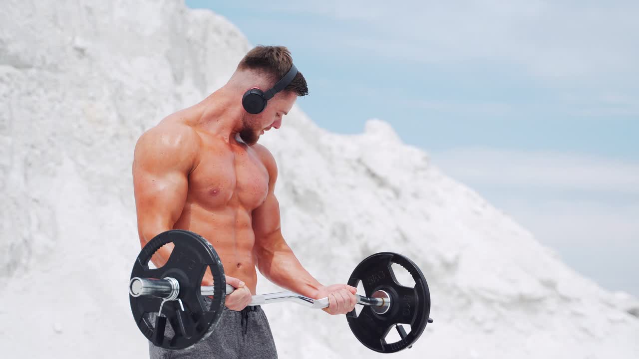 A man in headphones does barbell biceps exercises. Workout in nature