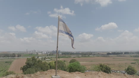 An-Israeli-flag-on-a-mountain-in-the-center-of-the-country,-with-buildings-and-roads-in-the-background-under-a-blue-sky-with-white-clouds