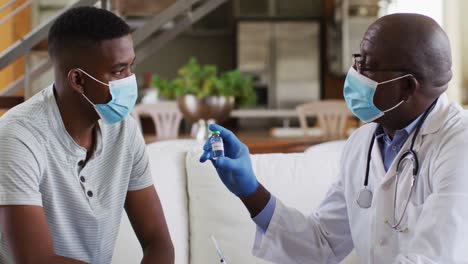 African-american-senior-male-doctor-giving-covid-vaccine-to-male-patient-in-home,-wearing-face-masks