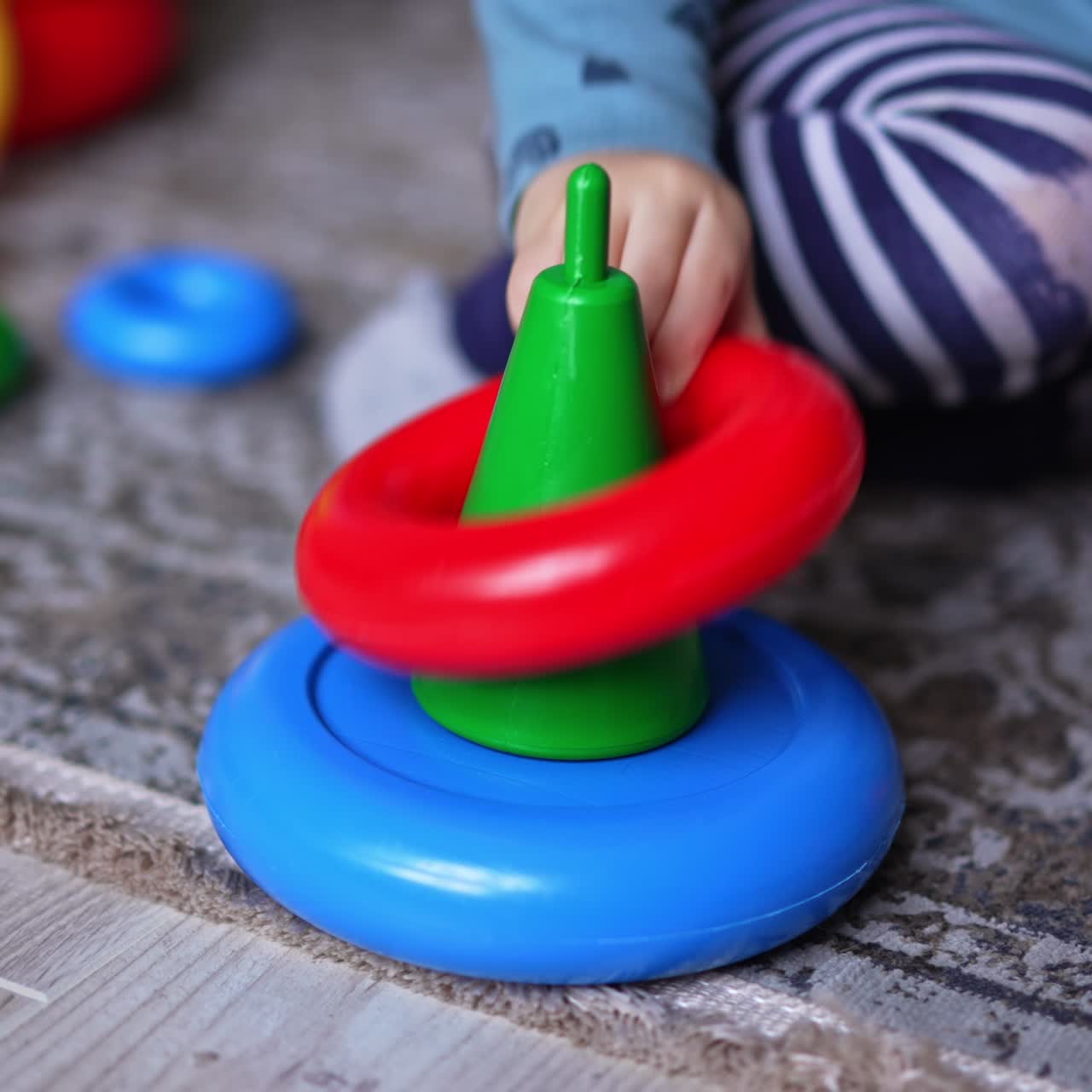 Little bright toy pyramid without upper parts. Close up. Unrecognized baby sitting on the floor next to a toy takes a red ring away. Blurred backdrop