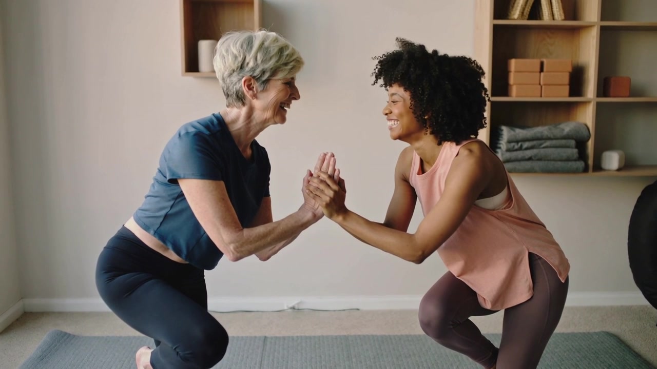 Pressing palms, two women lowering into one-leg squats at home balancing on gray mat in gymwear