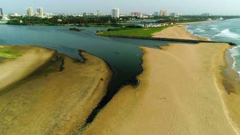 Drohnenaufnahmen-Eines-Strandes-Mit-Einem-Sich-öffnenden-Stauwasser-Und-Einer-Skyline-Der-Stadt-Mit-Hochhäusern-Im-Hintergrund