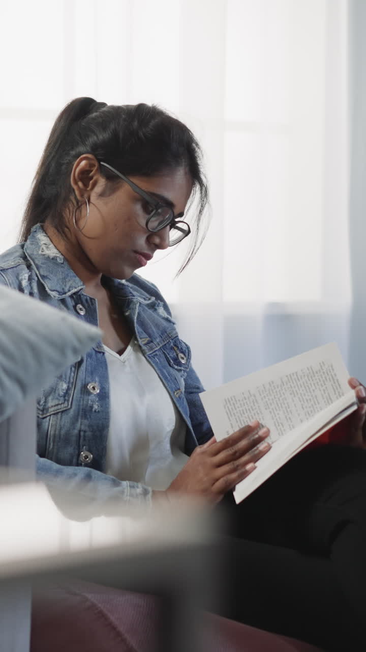 Attentive Indian student learns new information in textbook sitting in room against window. Brunette woman gets prepared for exams at home closeup