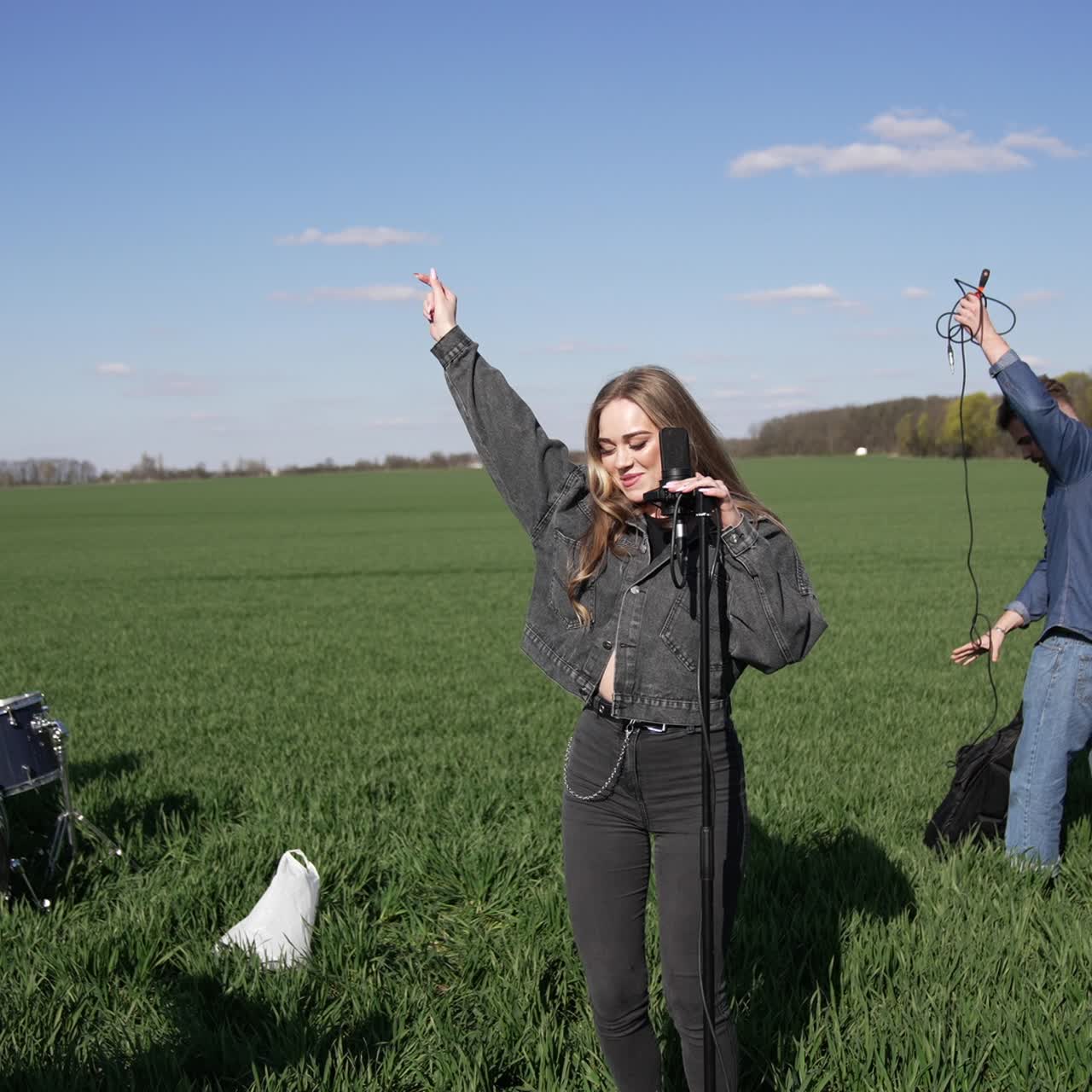 Energetic and cheerful musicians perform music on green field. Young band singing and dancing outdoors. Men and a woman play drums and sing a song into a microphone