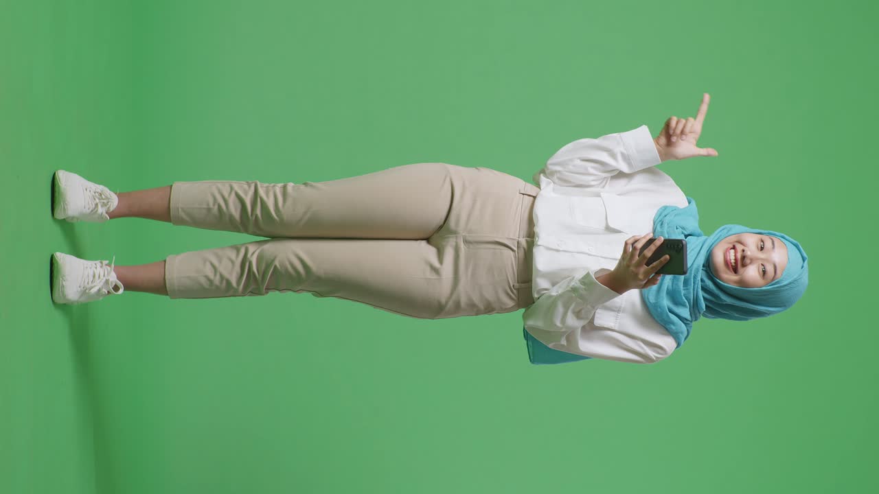 아시아계 무슬림 여성이 미소 짓고 스마트폰을 사용하다가 녹색 스크린 배경 스튜디오에 서서 을 가리키고 있다.