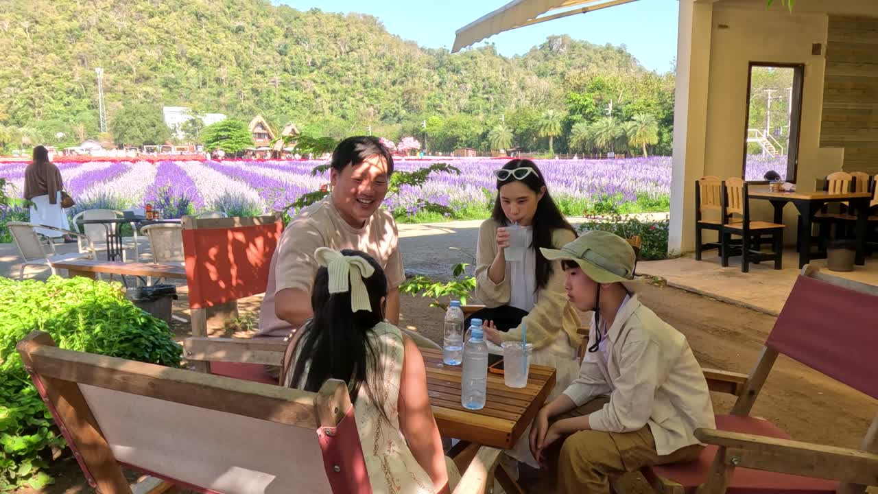 ครอบครัวชาวเอเชียที่มีความสุขกำลังพูดคุยและดื่มน้ำที่ร้านกาแฟกลางแจ้งข้างทุ่งลาเวนเดอร์สีม่วง