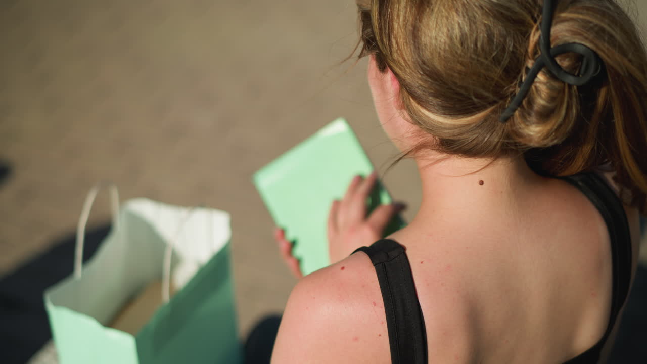 야외 벤치에 앉아있는 여자의 뒷면은 민트 녹색 가방에서 책을 가져오고 있습니다. 약간 흐릿합니다. 그녀는 책을 열고 페이지를 뒤집고 바닥에서 보이는 그림자를 가지고 있습니다.