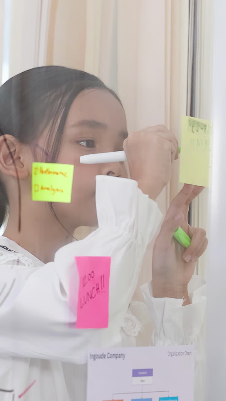 Young girl in white shirt writes on sticky notes on glass wall during brainstorming session