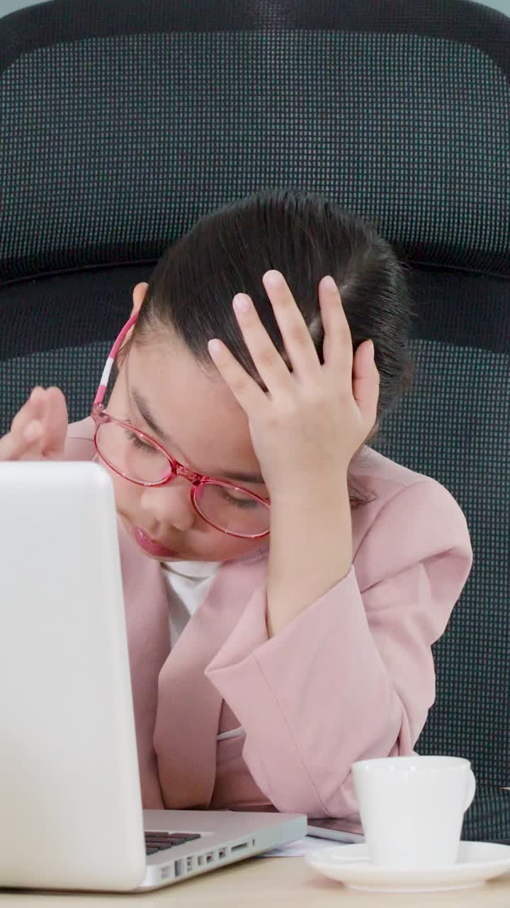 Una donna professionista esausta con un blazer rosa si tiene la testa mentre lavora al laptop.