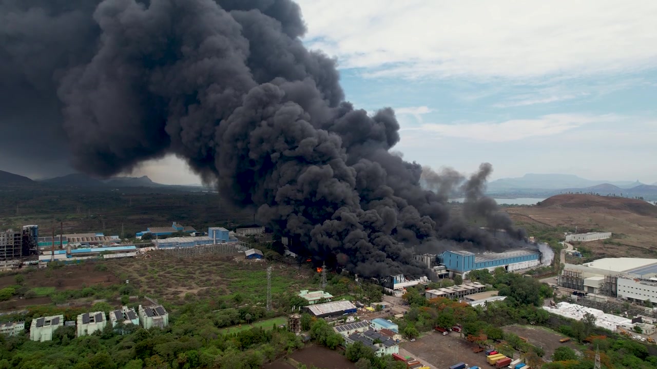 Vue aérienne large par drone d'un incendie industriel massif dégageant une épaisse fumée noire au-dessus d'un complexe industriel et du paysage environnant.