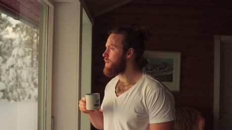 bearded caucasian guy is drinking coffee inside the winter cottage