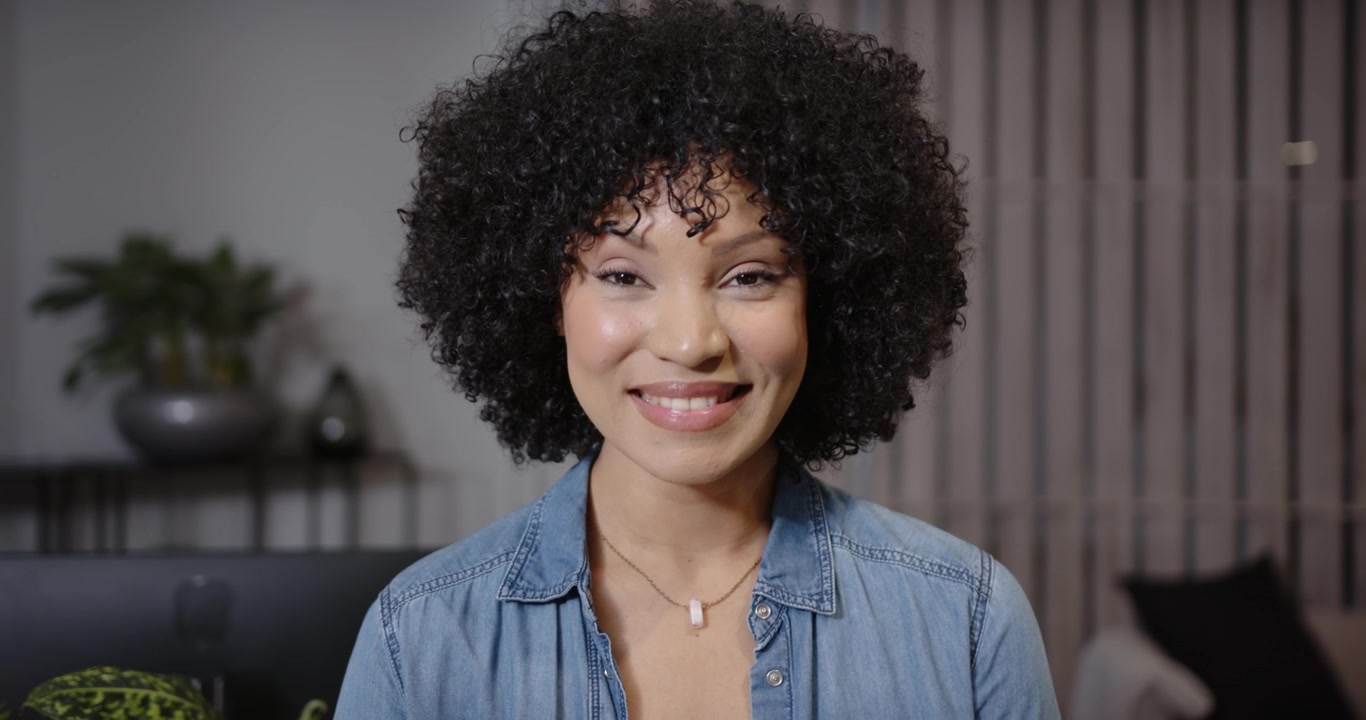 Donna afroamericana che guarda in alto verso la fotocamera, parla, sorride e ride a casa, con camicia di jeans e ciondolo