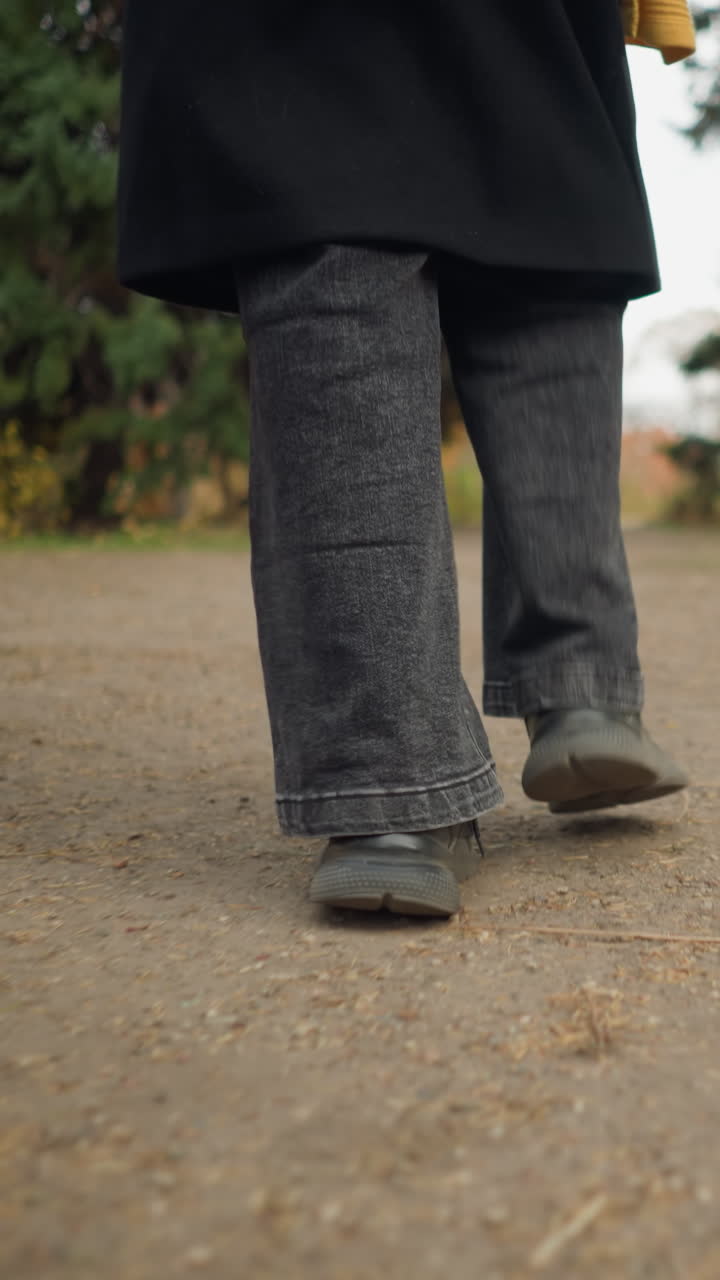 Close-up back view of feet in black jeans and canvas shoes walking along an autumn garden path, surrounded by colorful foliage and peaceful nature