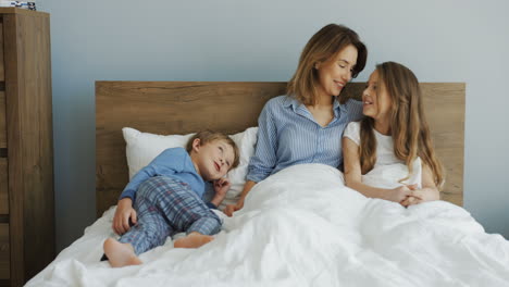 young beautiful mother sitting on the bed under the blanket and talking with her son and daughter in the morning